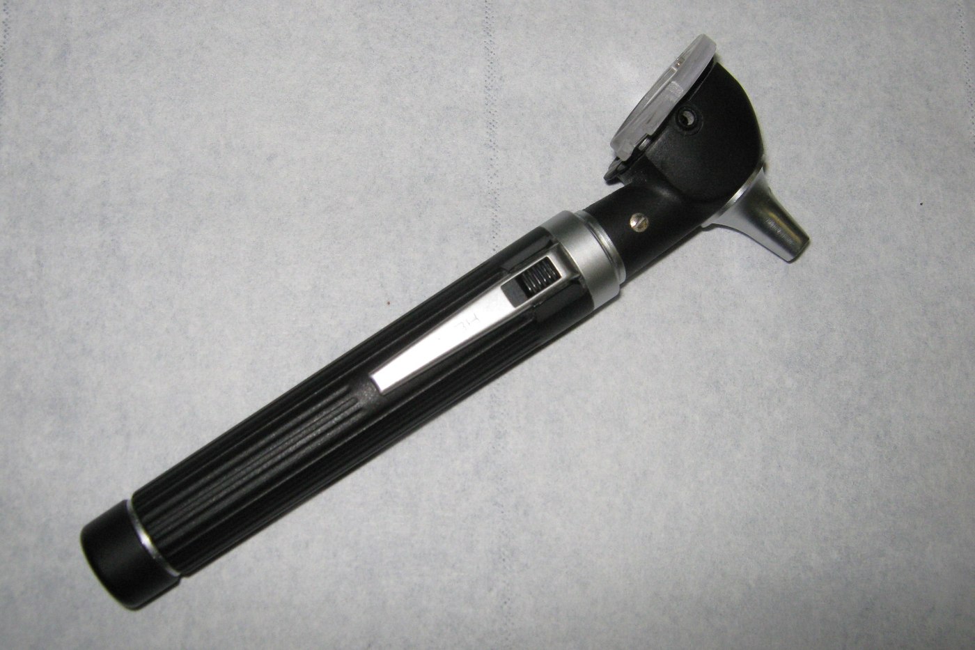 A black-and-silver otoscope rests diagonally on a pale examination-table covering. Illustrative photo.