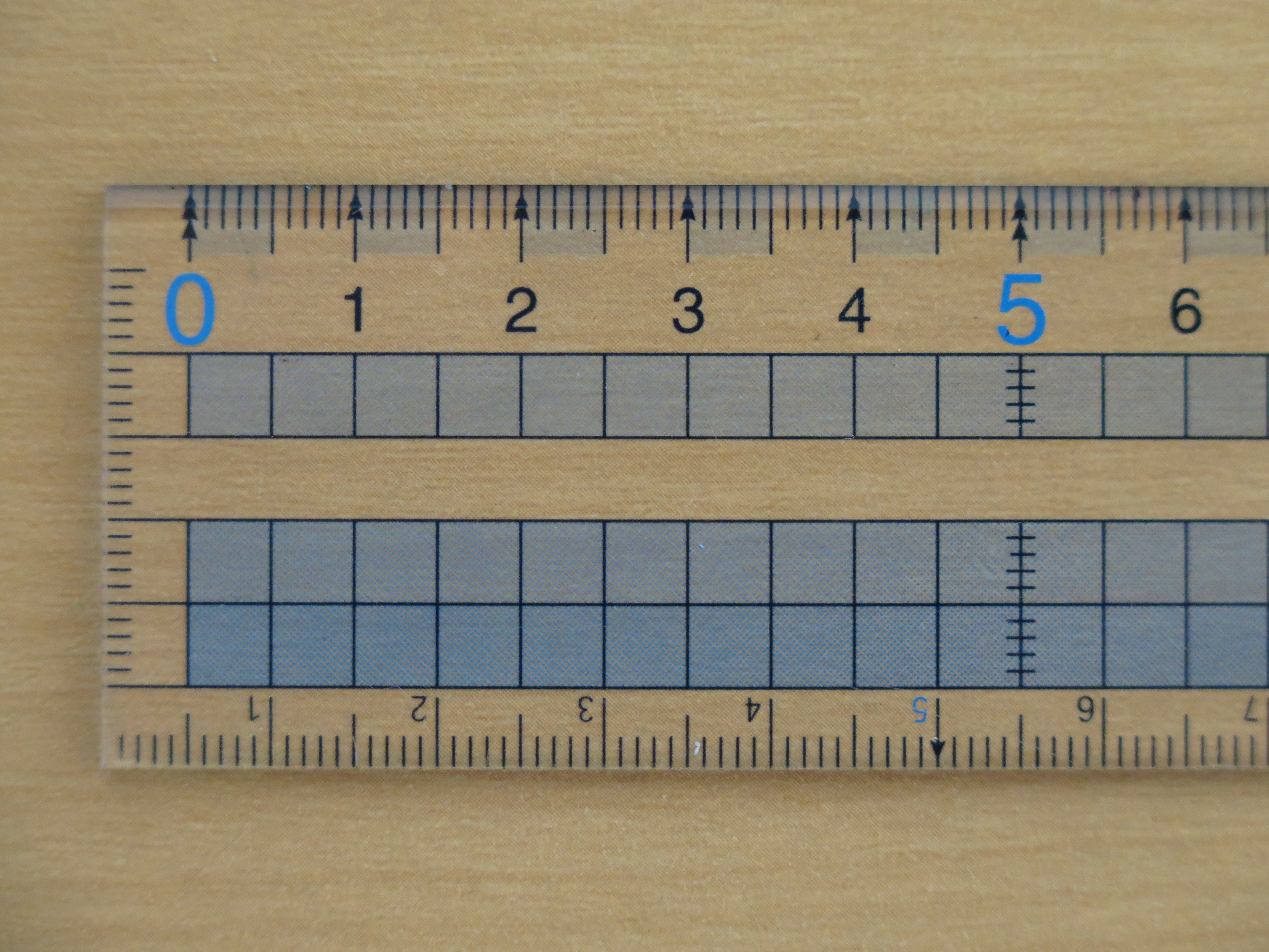 Closeup of a transparent ruler with blue and black centimeter markings on a wooden tabletop. Illustrative photo.