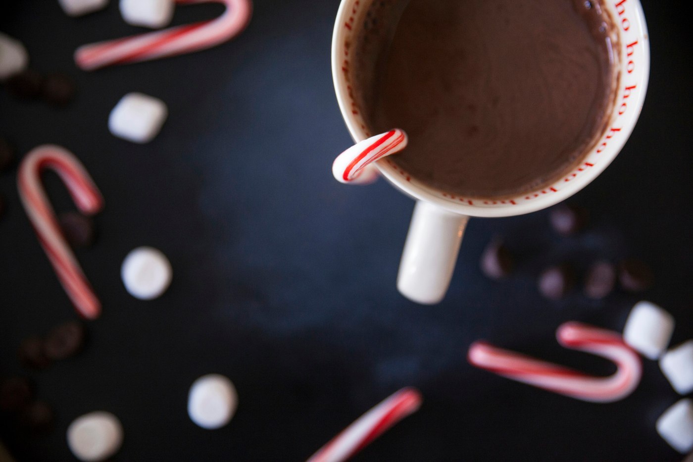 A white mug of hot cocoa with a candy cane sits on a dark surface among candy canes, marshmallows and chocolate pieces. Illustrative photo.