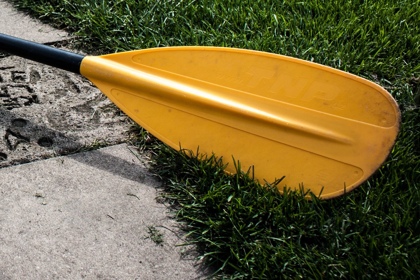 Closeup of a yellow kayak paddle blade and black shaft resting beside gray paving and green grass. Illustrative photo.