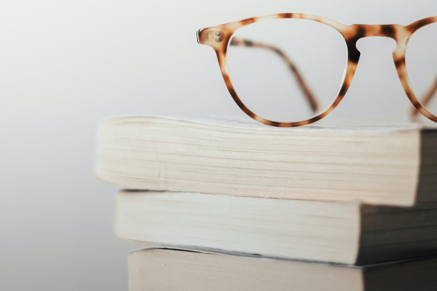 Tortoiseshell reading glasses rest on the pale page edges of a stack of books against a plain light background. Illustrative photo.