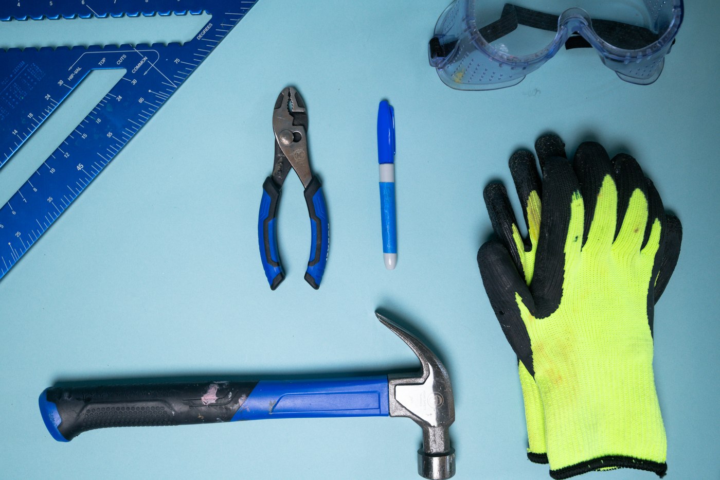 A claw hammer, pliers, carpenter's square, marker, safety goggles and work gloves arranged on a light blue surface. Illustrative photo.