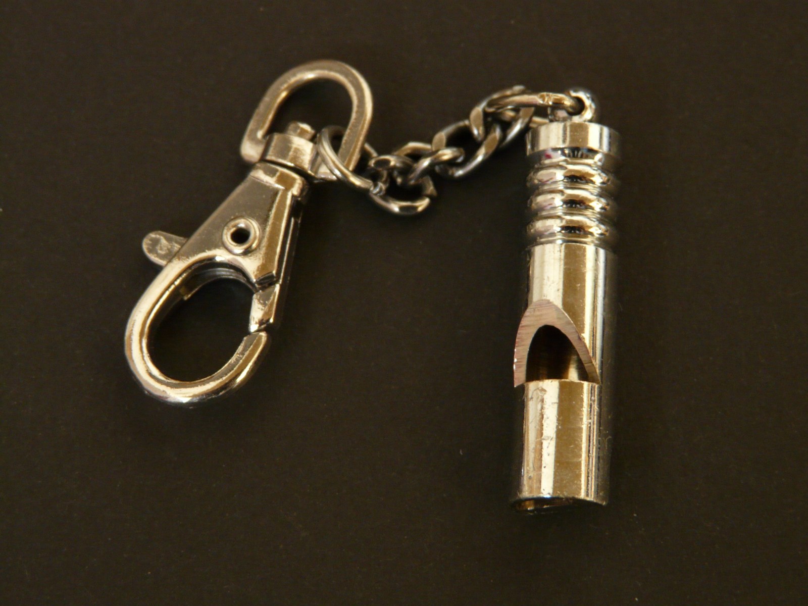Closeup of a silver metal whistle attached to a short chain and clasp on a dark surface. Illustrative photo.